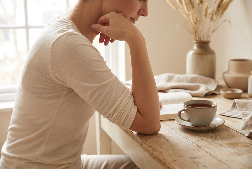 Frau in cremefarbenem Langarmshirt stützt sich beim Lesen auf den angewinkelten Arm, daneben eine Tasse Tee