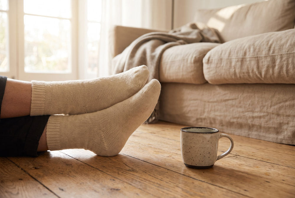 Füße in hellen Socken liegen übereinander auf einem Holzboden vor einem beigen Sofa, daneben eine Tasse